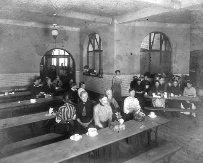 Victorian Edwardian pictures of women seated in a communal dining hall with wooden benches and cups on tables.