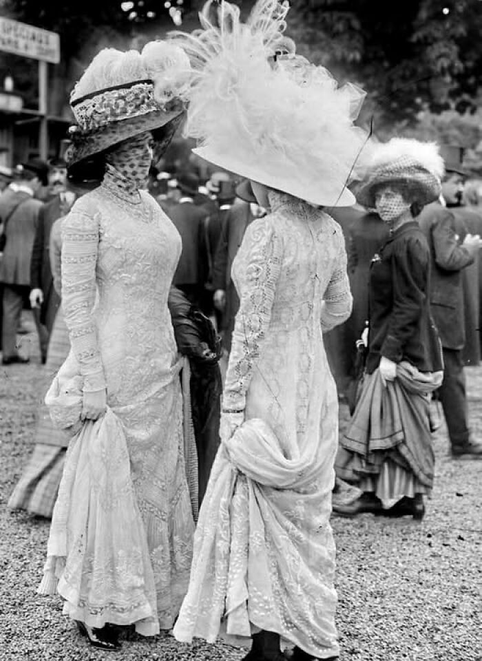 Two women dressed in intricate Victorian Edwardian pictures style gowns and elaborate hats at an outdoor event.