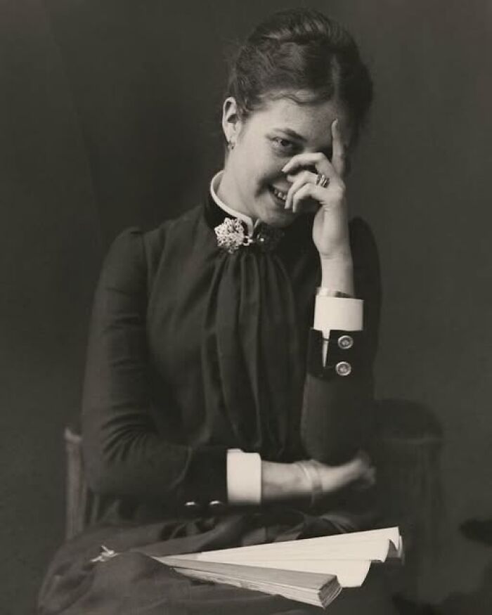 Young woman in Victorian-Edwardian clothing smiling shyly while seated with a folded fan in her lap in vintage style.
