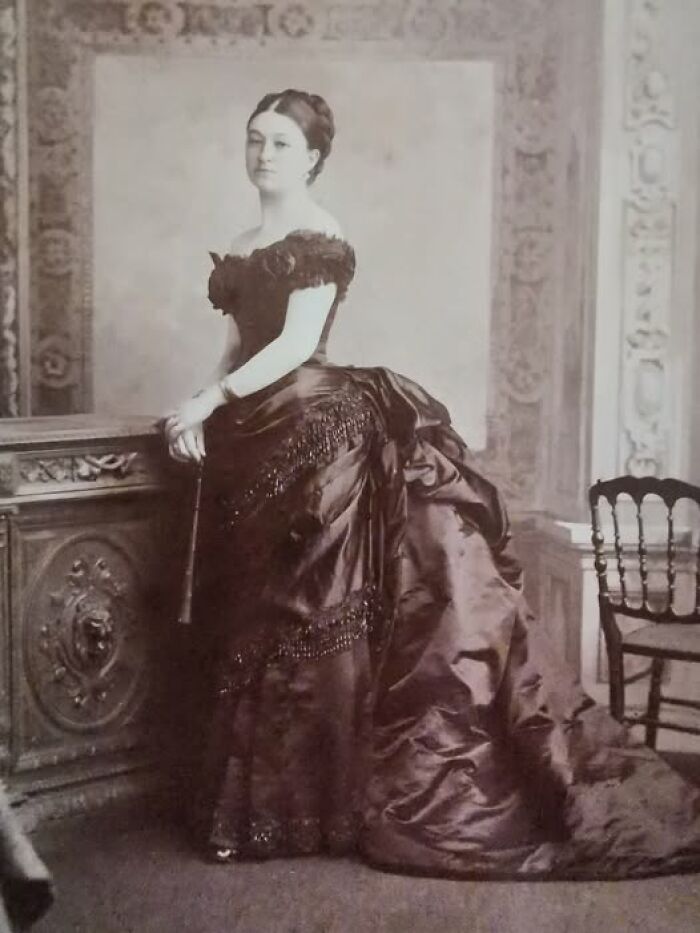 Victorian-Edwardian pictures of a woman in an ornate dark dress standing beside an intricately carved wooden piece of furniture.