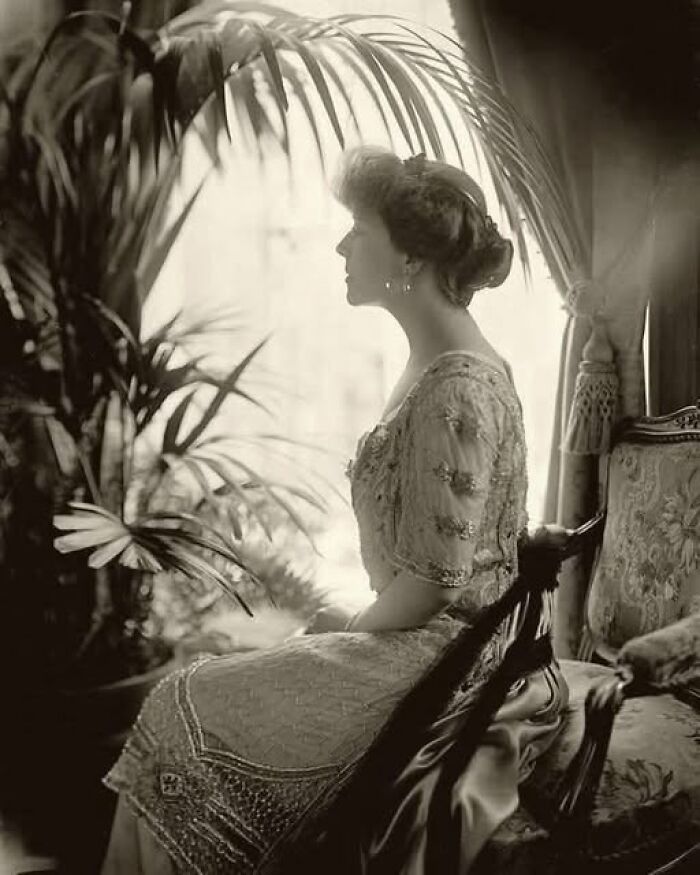 Victorian-Edwardian picture of a woman in ornate dress seated near a window with decorative plants and antique furniture.