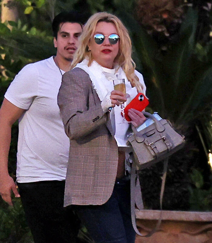 Britney Spears looks unrecognizable wearing sunglasses and holding a drink and purse while leaving a bar with a man behind her.