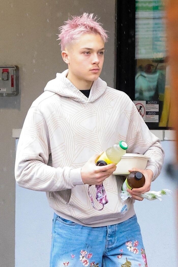 Knox Jolie-Pitt with pink hair, wearing a patterned hoodie and floral jeans, holding drinks and a bowl outdoors.