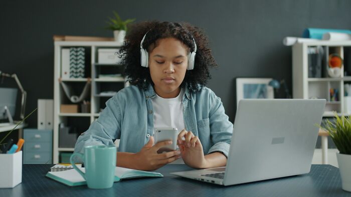 Young Gen-Z worker wearing headphones, using a smartphone and laptop at a desk in a modern office setting.