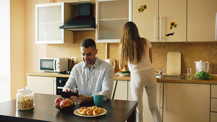 Man relaxing with tablet while woman does domestic chores in kitchen, highlighting unequal household responsibilities.