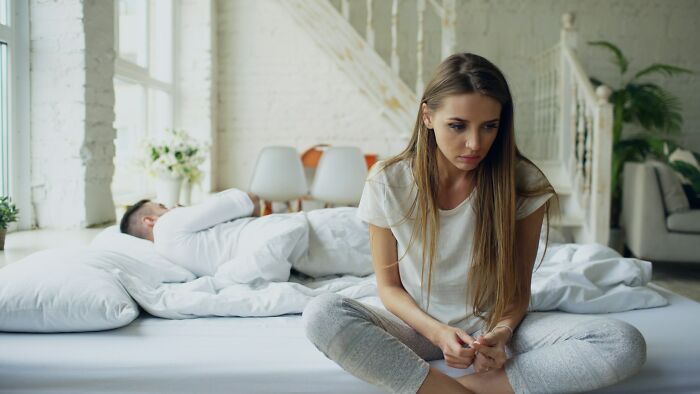 Young woman sitting on bed looking upset while man lies asleep in the background, showing subtle signs of cheating behavior