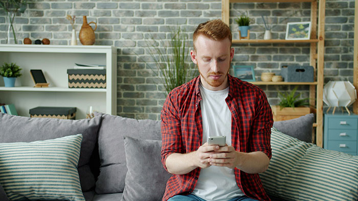 Young man in red plaid shirt sitting on couch distracted by phone, ignoring domestic chores and wife&rsquo;s expectations at home.