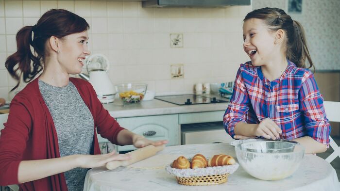 Mother and daughter laughing while rolling dough together in the kitchen, showing kids doing something clearly passed down from parents.
