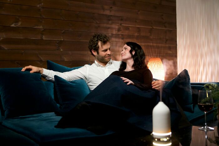 Couple sitting closely on a dark blue couch in a cozy room, showing subtle signs that suggest they might be cheating.