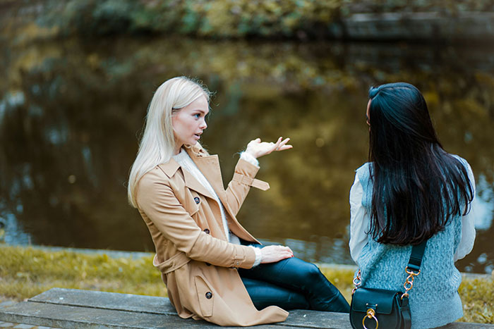 Two women sitting outdoors by water, one gesturing while discussing avid walker doing 20K steps per day.