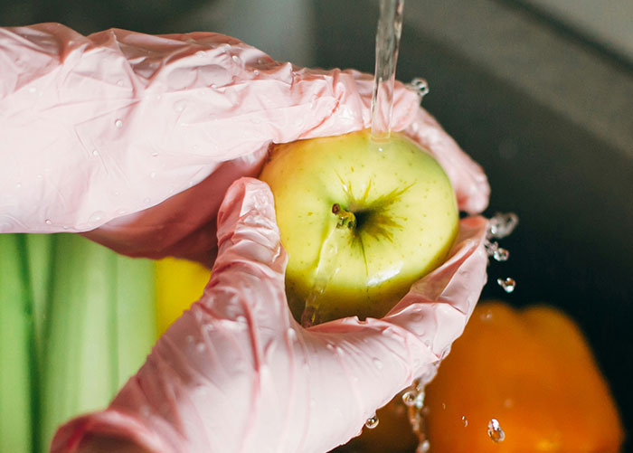 Washing fruits and vegetables under running water while wearing pink gloves to ensure cleanliness and safety.