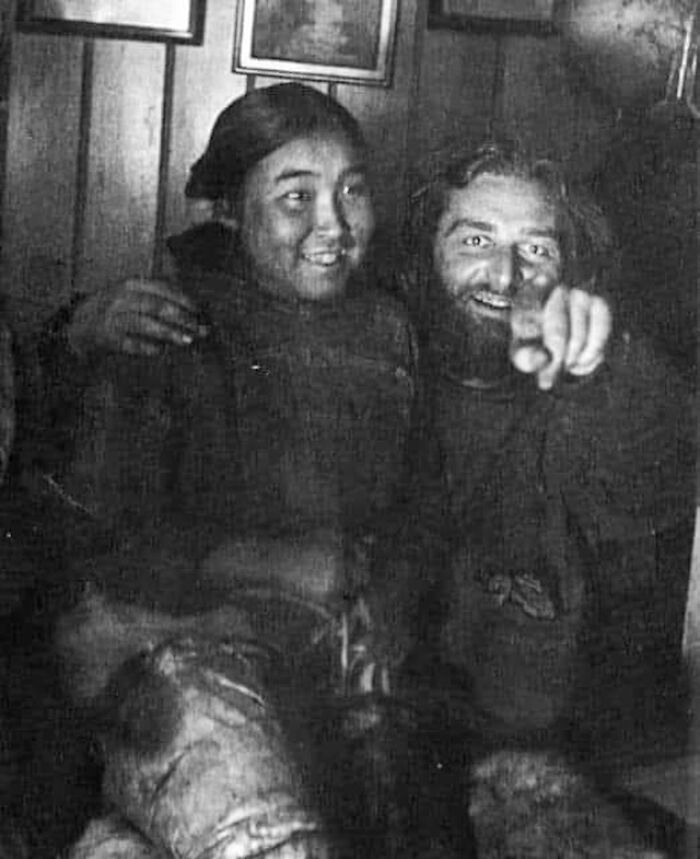 Victorian era weird photo showing two people smiling and pointing in a dimly lit room with framed pictures on the wall.