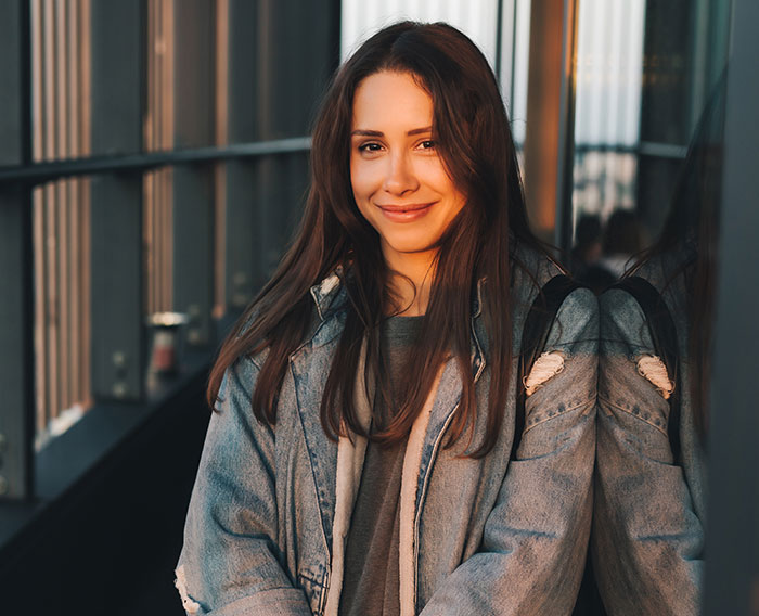 Young woman smiling in a denim jacket leaning against a reflective glass wall, representing unhinged women men avoid.