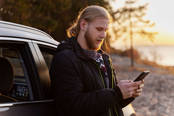 Young man standing by car on beach at sunset, looking at phone, illustrating men’s perspective on unhinged behaviors.