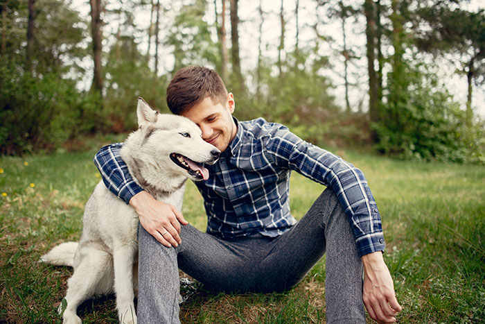 Man sitting on grass outdoors, hugging a happy husky dog, enjoying a peaceful moment in nature.