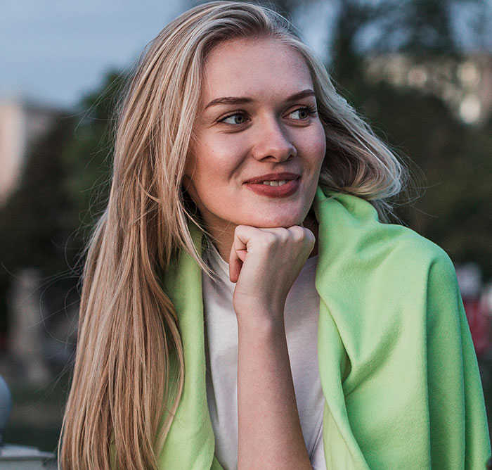 Young woman with long blonde hair and green shawl, smiling thoughtfully outdoors, representing women who did unhinged things.