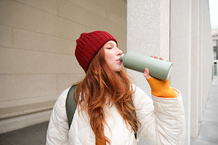 Young woman with red hair wearing a beanie and coat drinking from a reusable bottle showing independent women behavior.