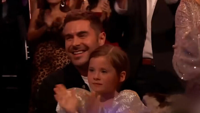 Zac Efron smiling and clapping with a child at an event, drawing attention to his back to normal face appearance.