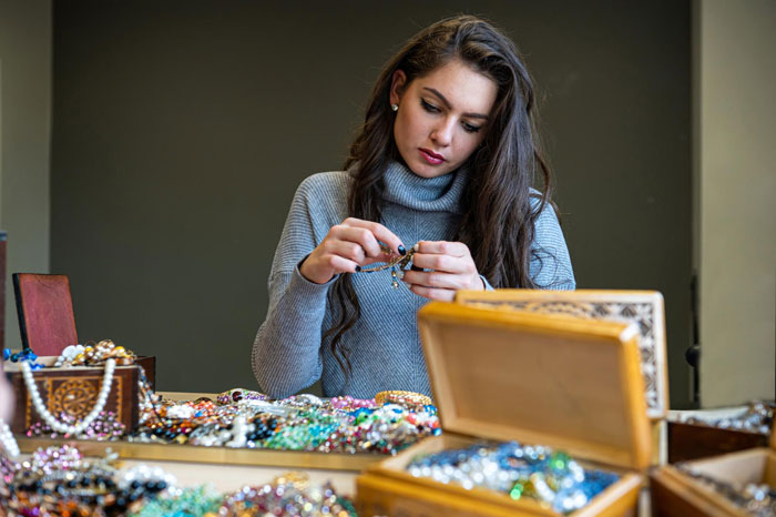 Woman Etsy artist examining bracelets at a table filled with handmade jewelry and decorative boxes in a cozy room. Woman Etsy artist examining bracelets at a table filled with handmade jewelry and decorative boxes in a cozy room.