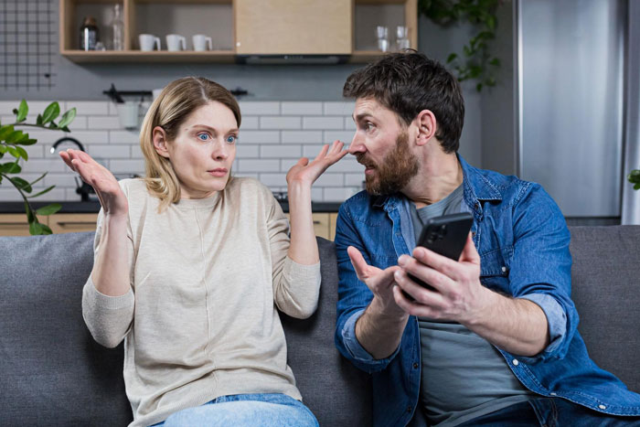 Couple having a tense conversation on couch during family vacation, husband focused on business, wife looks frustrated and conflicted.