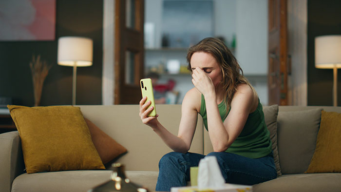 Woman sitting on couch looking distressed while holding phone, reflecting on suspicious work trip and relationship doubts.