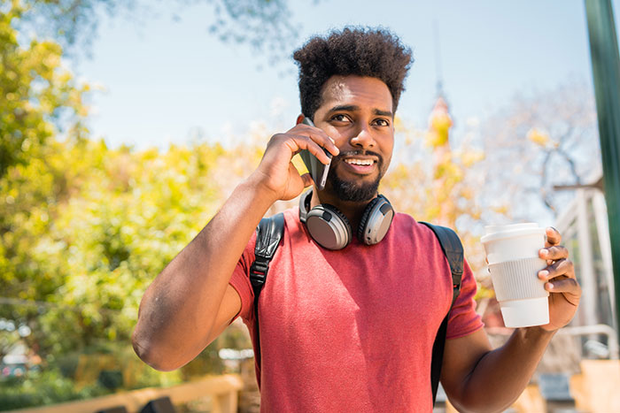 Man wearing headphones and red shirt, holding coffee and talking on phone during suspicious work trip outdoors.