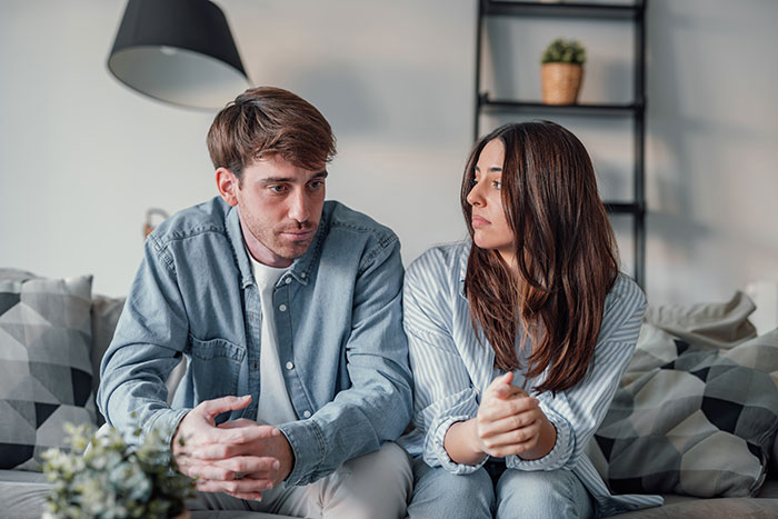 Young couple sitting on couch looking upset, depicting tension over guy secretly giving his sister a key to GF&rsquo;s house.