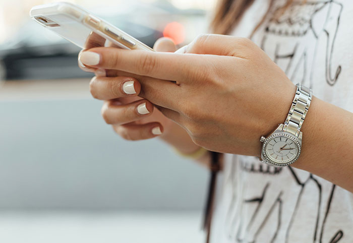 Woman child holding smartphone with manicured nails and silver watch, wearing a casual graphic t-shirt outdoors.