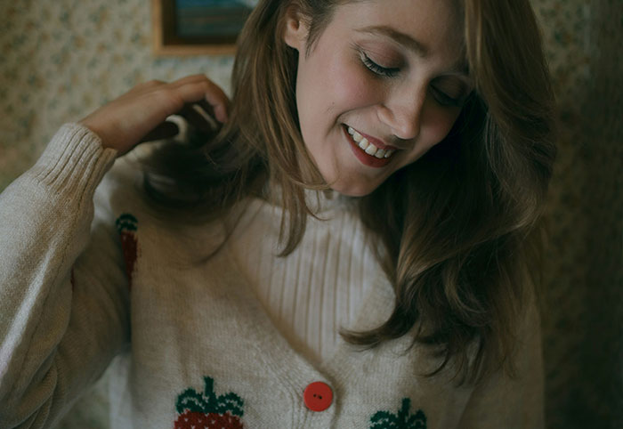 Young woman smiling gently, wearing a cozy sweater with strawberry patterns, reflecting a woman child vibe.