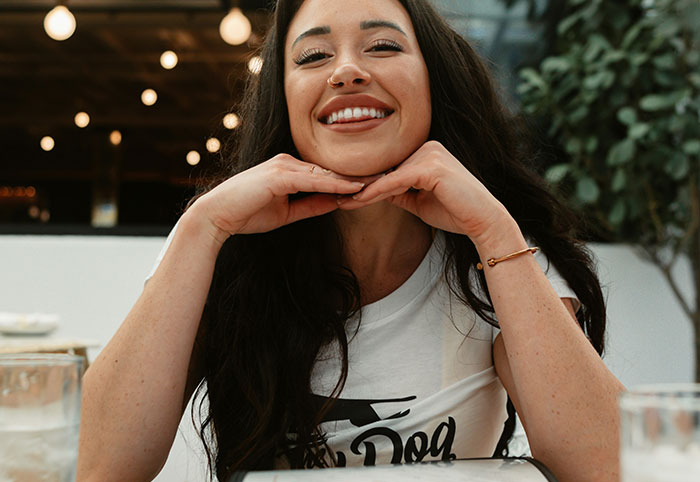 Smiling woman with long dark hair resting her chin on hands, expressing joyful woman child energy in casual setting