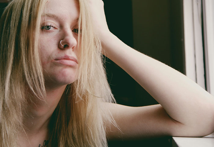 Young woman child with long blonde hair and nose ring resting head on hand by a window in natural light.