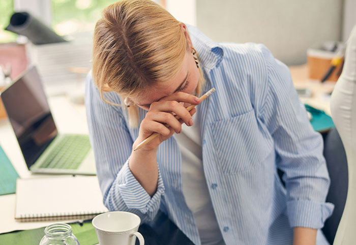 Young woman in casual shirt covering her face with hand, showing signs of stress or overwhelm in a workspace setting