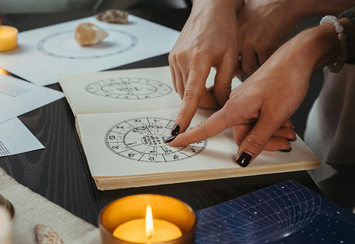 Two women pointing at astrological charts in a book, surrounded by crystals and a lit candle, woman child vibe.