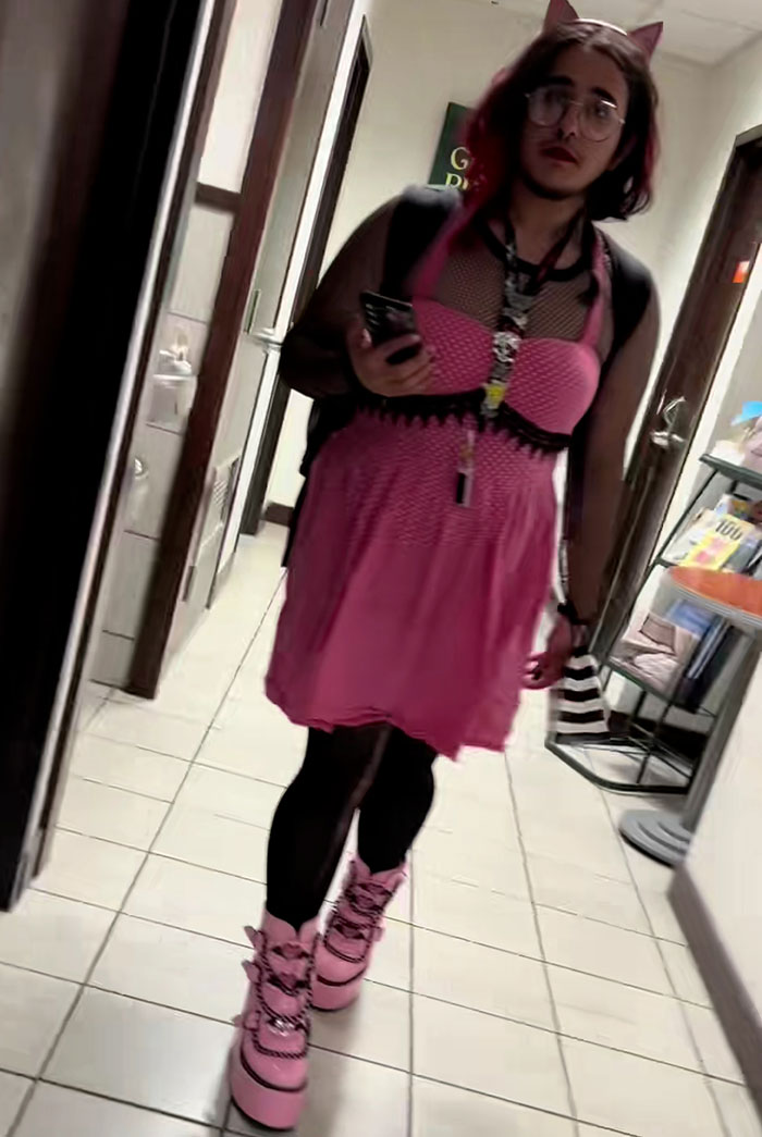 Bearded trans man wearing pink dress and platform shoes walking down hallway, related to restroom debate controversy.