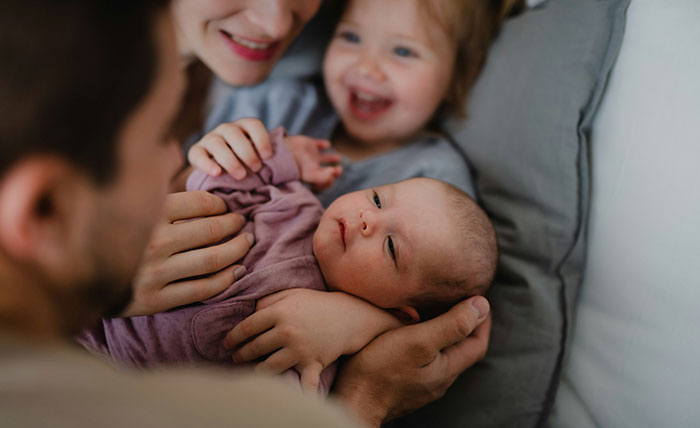 Mother and father holding a newborn baby while toddler smiles, highlighting favorite grandchild and family tension.