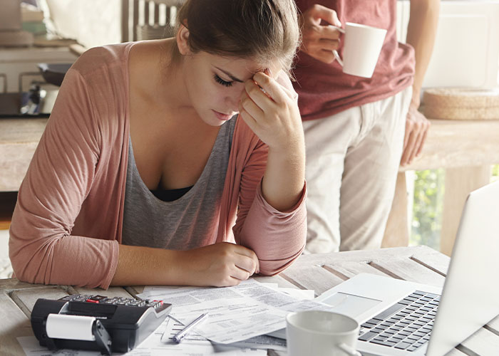 Stressed woman reviewing bills at table while partner stands behind her with a coffee, reflecting jobless partner burdens.