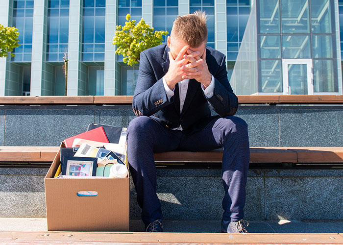 Man in suit sitting on steps with head in hands next to box of personal items after job loss or breakup.