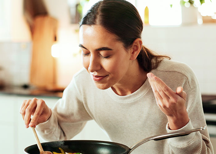 Woman cooking in a kitchen, tasting food from a pan, showing reluctance toward foreign traditional Christmas spread.