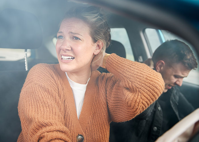 Upset couple inside a car after crash, woman wearing brown sweater showing distress, man in black jacket seated behind.