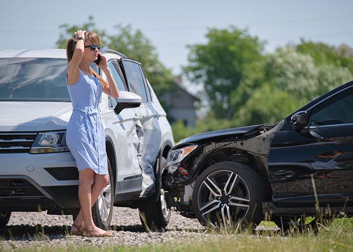 Woman in blue dress making phone call beside damaged cars after accident, reflecting upset feelings in a car crash scenario.