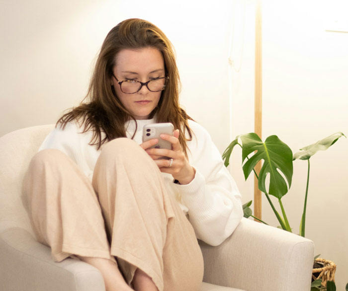 Woman in glasses sitting thoughtfully on chair, focusing on her phone, reflecting on traditional family Thanksgiving hosting.