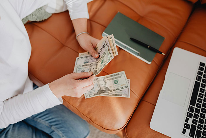 Woman counting cash on a couch next to a laptop and notebook, considering a $4k plastic surgery request.