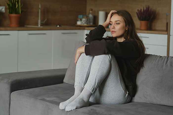 Young woman sitting on couch looking thoughtful, contemplating sister&rsquo;s $4k plastic surgery and body dysmorphia concerns.