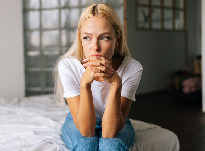 Woman refusing homeless sister-in-law to move in, sitting thoughtfully on bed in a casual white shirt and jeans.
