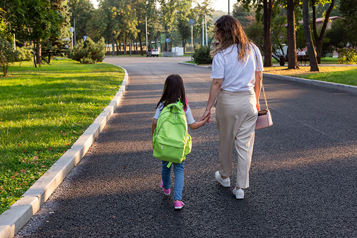 Woman holding niece's hand walking to school on quiet road surrounded by trees in early morning light