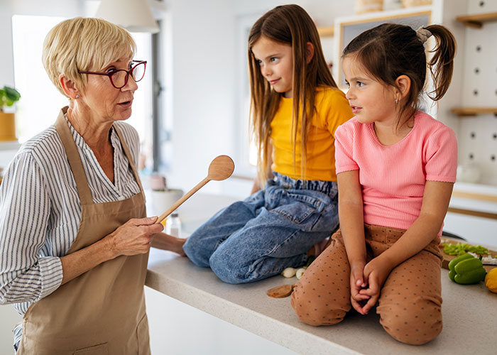 Grandma babysitting two kids in the kitchen, holding a wooden spoon and talking to them while they sit on the counter. Grandma babysitting two kids in the kitchen, holding a wooden spoon and talking to them while they sit on the counter.