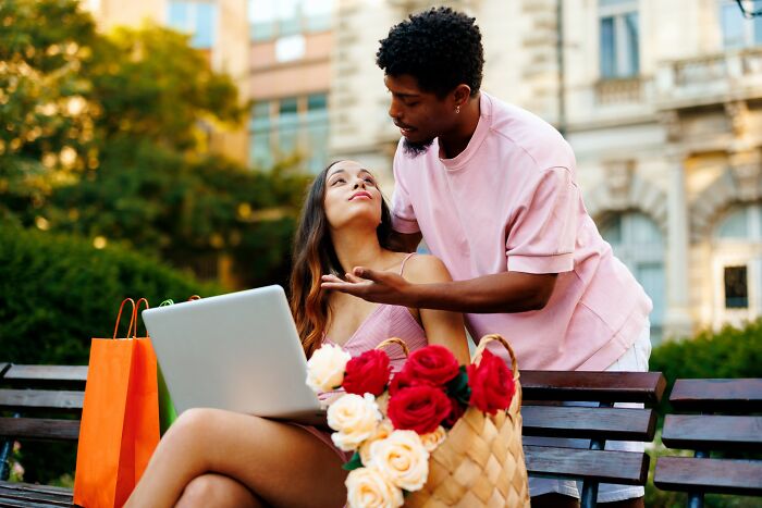 Young couple on park bench with laptop and shopping bags, discussing a situation after falling for scams online.