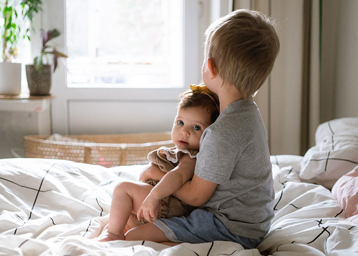 Young teen boy sitting on bed holding baby sibling, illustrating babysitting step-siblings and family tension themes.
