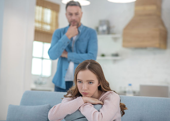 Teen girl upset on couch while dad stands in background looking frustrated in a modern home living room.