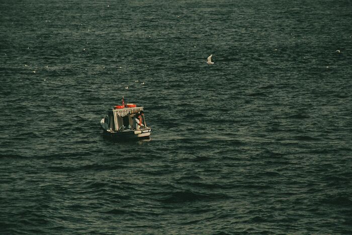 Small boat floating on vast dark water with a lone person creating a bizarre and surreal moment at sea.